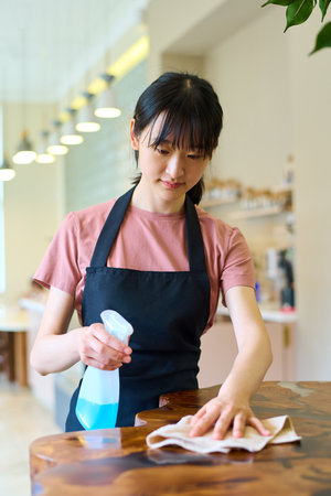 Young Asian waitress in apron wiping table with cleaning agent before opening of cafeの写真素材