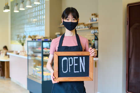 Asian waitress in face mask standing at coffee shop holding open sign during  pandemic