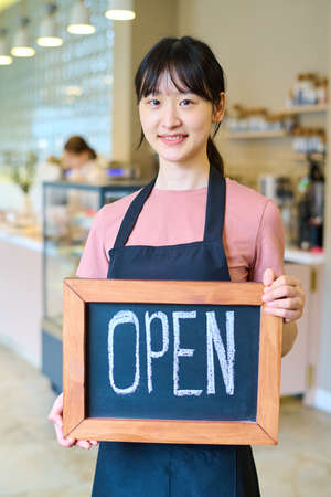 Portrait of young Asian waitress in uniform holding sign and smiling at camera, she opening cafe