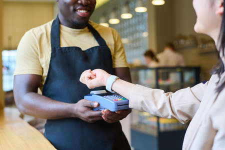 Close-up of woman using her smartwatch to pay online for order while waiter holding terminal