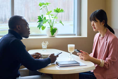 Two business partners exchanging phone numbers while drinking coffee at table during meeting at cafe