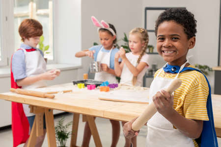 Hands of child with wooden pin rolling fresh dough on table while helping mom with cooking pastryの写真素材
