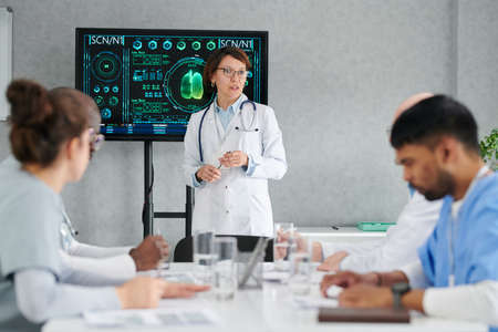 Mature female woman standing near the screen with charts and talking to her colleagues during...