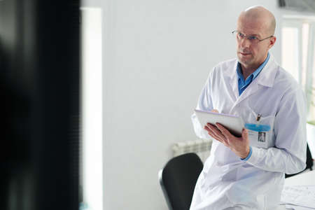 Serious mature male doctor in white coat using digital tablet during online conference with colleaguesの写真素材
