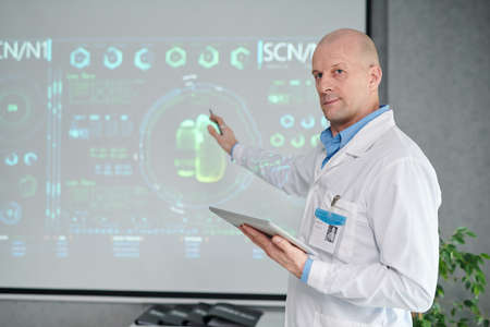 Portrait of mature male doctor in white coat with digital tablet pointing at projector with medical...