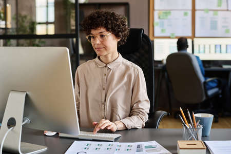 Young businesswoman in eyeglasses sitting at her workplace in front of computer monitor and typing