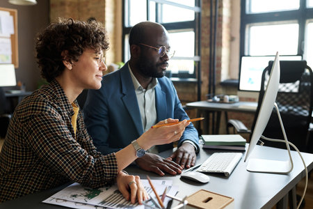 Two multiethnic colleagues sitting at table in front of computer monitor and having online...