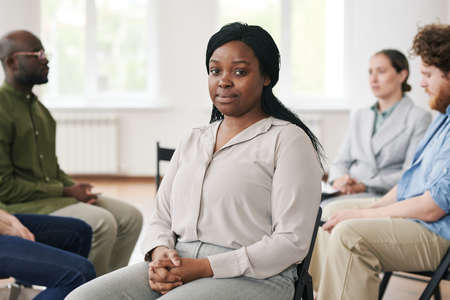 Young black woman in casual white blouse and pants sitting in front of camera and looking at you against group of people having sessionの写真素材