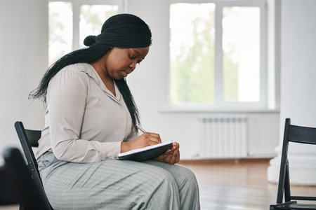 Young serious black woman making notes in notebook before or after session of psychotherapy while...