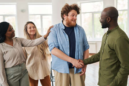 Two young happy interracial men shaking hands before or after psychological session while standing against cheerful femalesの写真素材