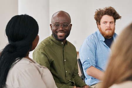 Happy young black man looking at one of patients during psychological session while sitting among other people and listening to herの写真素材