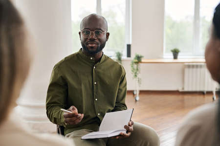 Serious young male psychologist with notebook and pen consulting two intercultural female patients...