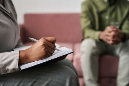 Hand of young female psychoanalyst making notes in notebook during session while listening to patient describing his worriesの写真素材