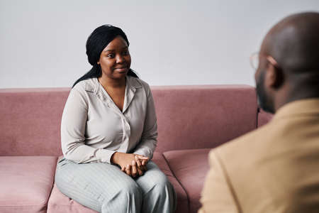 Young smiling African American female patient looking at psychoanalyst giving her advice while sitting on couch during psychological sessionの写真素材