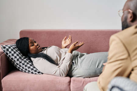 Young stressed female patient lying on couch and describing her problem to psychoanalyst sitting in front of her and giving adviceの写真素材