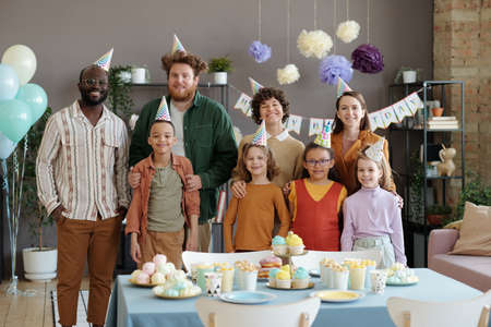 Portrait of happy adults in party hats smiling at camera together with their children standing at table with sweet food during birthday partyの写真素材