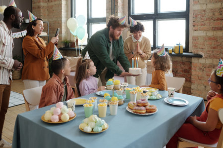 Young father holding birthday cake while his son blowing candles on it and guests shooting video on...