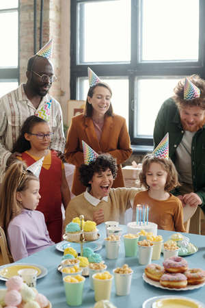 Little boy in party hat blowing candles on birthday cake while sitting at table with dessert and celebrating birthday with adults and childrenの写真素材