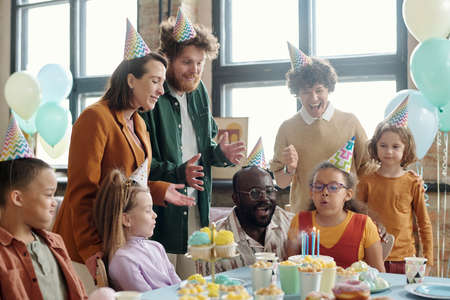 Little girl in party hat blowing candles on birthday cake with her parent and friends congratulating her with birthdayの写真素材