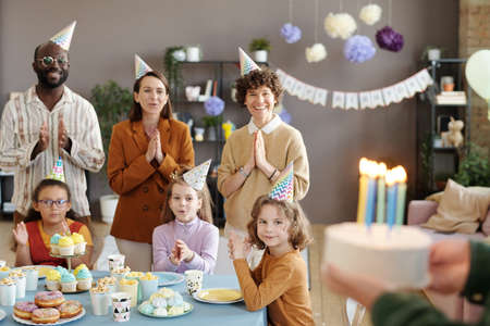 Group of happy people in party hats standing and waiting for birthday cake together with children sitting at table with dessertの写真素材