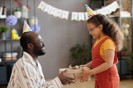 African American father in party hat happy to congratulate his daughter with her birthday giving gift box to herの写真素材