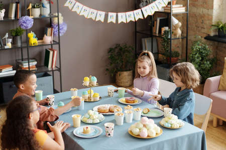 Group of children playing rock paper scissors game while sitting at table with sweet food in living room at birthday partyの写真素材