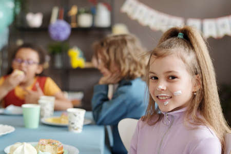 Portrait of little girl with cream on her face smiling at camera while sitting at table with her friends and eating dessert at partyの写真素材