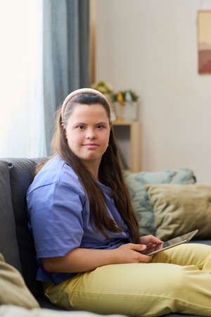 Cute girl with disability in casualwear using tablet at leisure while sitting on sofa in home...