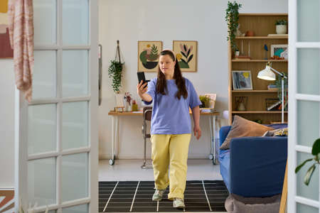 Girl with Down syndrome in casualwear holding smartphone in front of herself while moving along...