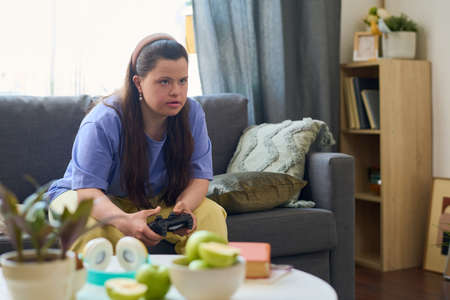 Girl with Down syndrome in casualwear looking at tv screen while sitting on couch and playing video...