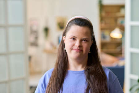 Young happy female with Down syndrome standing in front of camera in living room and looking at you...