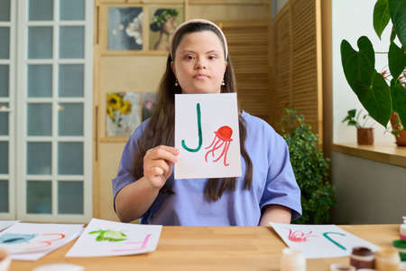 Young serious teacher showing paper with drawn green letter J and red jellyfish while sitting by...