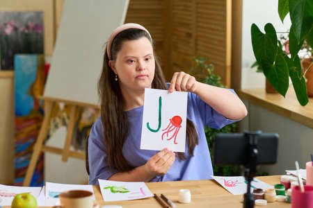 Girl with Down syndrome showing paper with English letter and drawing of jellyfish to online audience while sitting in front of cameraの写真素材