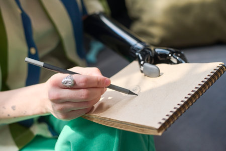 Hand of young woman with physical disability holding pencil over blank page of notepad while sitting...