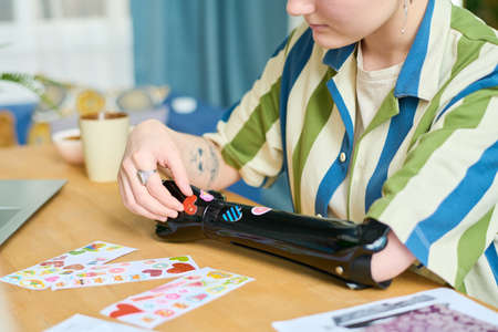 Young female with myoelectric arm sitting by table in living room and putting small handmade...