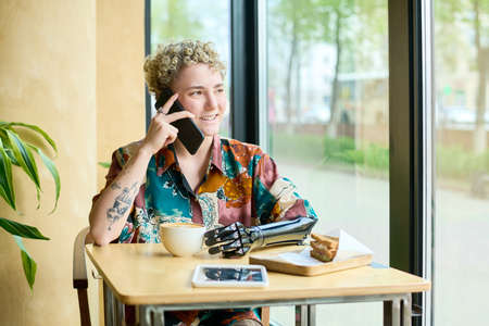 Happy young female with partial arm speaking on mobile phone while sitting by table in cafe, looking...