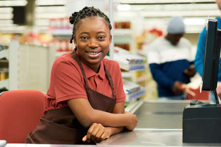 Happy young checkout staff in red t-shirt and brown apron sitting by workplace in modern supermarket and looking at cameraの写真素材