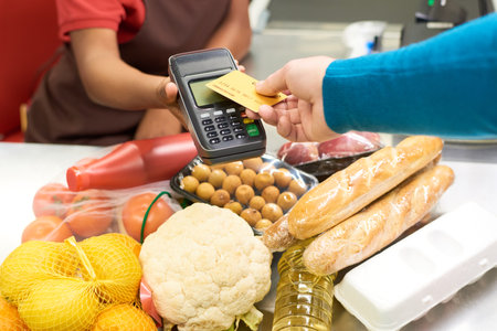 Hand of male consumer holding credit card over screen of payment terminal held by African American...