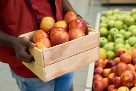 Hands of young black man holding wooden box with fresh ripe red apples while standing by large display with other fruits in supermarketの写真素材