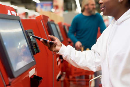 Young black woman with smartphone making contactless payment by terminal of self service while...