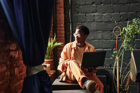 Young serious African American man with laptop sitting by windowsill and looking through window...