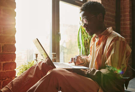 Serious guy in eyeglasses, casual shirt, pants and leather jacket holding laptop on his knees while...