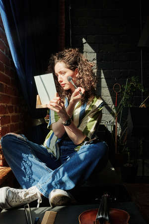 Brunette girl with wavy hair holding mirror in front of her face while applying make-up by window in...