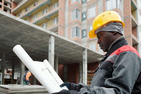 Serious mature black man in workwear looking at sketch of building structure while learning details of paper at construction siteの写真素材