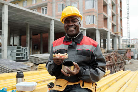 Happy mature black man with sandwich and cup of hot tea looking at camera while sitting on stack of...