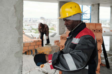 Mature male builder with electric drill carrying out his work in unfinished construction against colleague putting brick layer while building wallの写真素材
