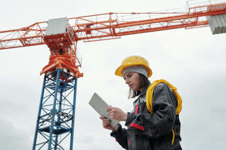 Young serious female engineer in workwear and protective helmet looking at screen of tablet while...