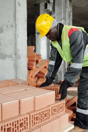 Mature experienced black man in workwear and hardhat bending over stack of bricks at construction...