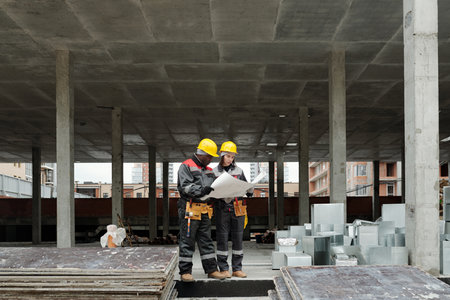 Small group of intercultural engineers or builders in workwear looking at plan of construction on...