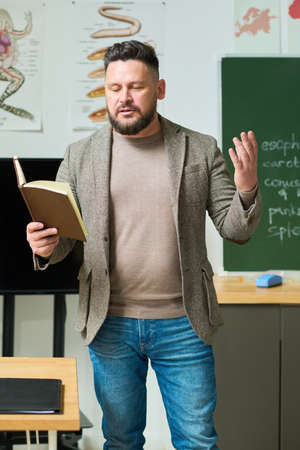 Confident professor or school teacher looking in open book while explaining his students new subject or making speech at lessonの写真素材